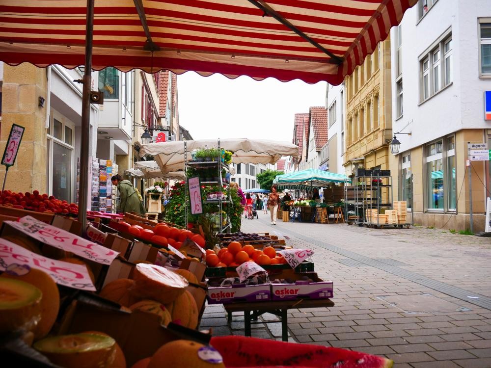 Marktstand mit Obst und Gemüse unter rot-weiß gestreiftem Sonnenschutz an einer gepflasterten Straße mit weiteren Marktständen und historischen Gebäuden im Hintergrund