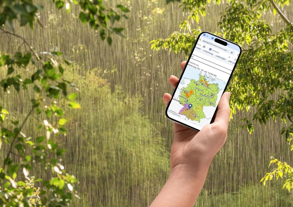 Symbolfoto: Eine Hand hält ein Handy auf dessen Display die Website des Deutschen Wetterdiensts zu sehen ist.  Im Hintergrund regnet es.