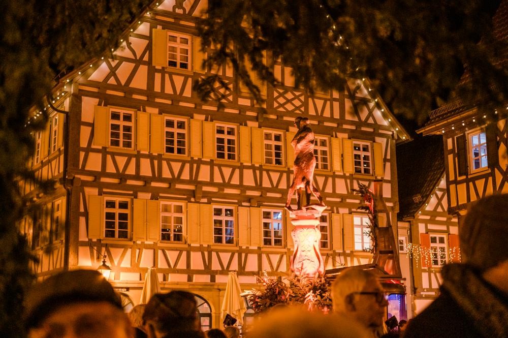 Beleuchteter Fachwerkhaus-Hintergrund mit einer Statue auf einem Sockel, davor eine Menschenmenge bei Nacht.