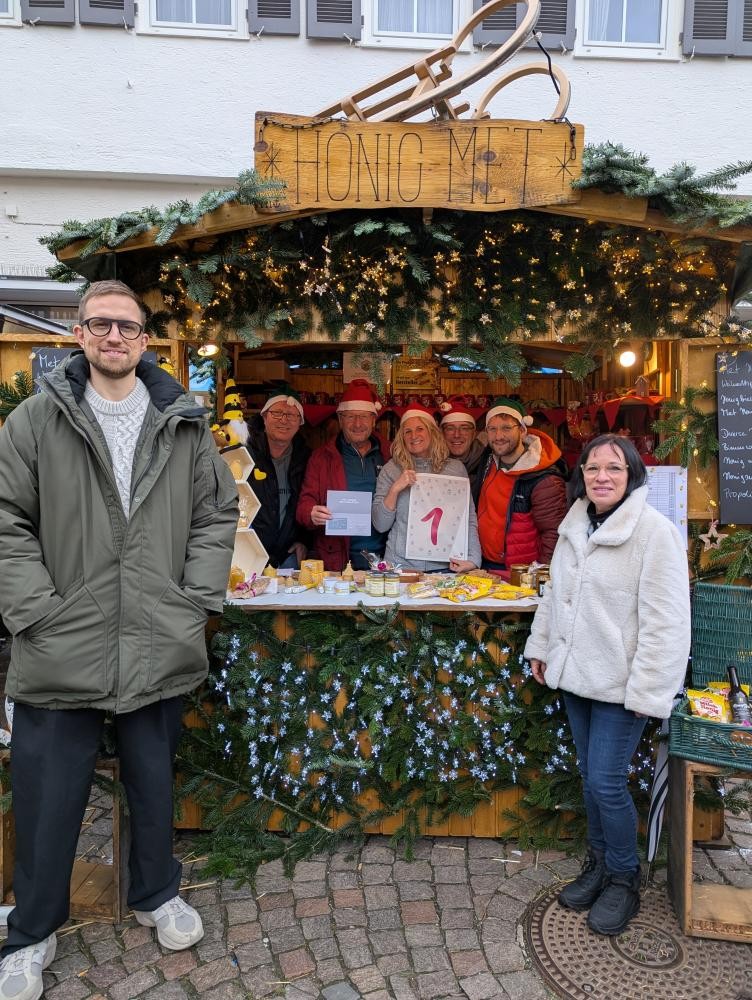 Weihnachtsmarktstand mit Tannengrün und Lichterketten, Holzschild 'HONIG MET', mehrere Personen hinter und vor dem Stand, eine Person hält ein Schild mit roter '1'.