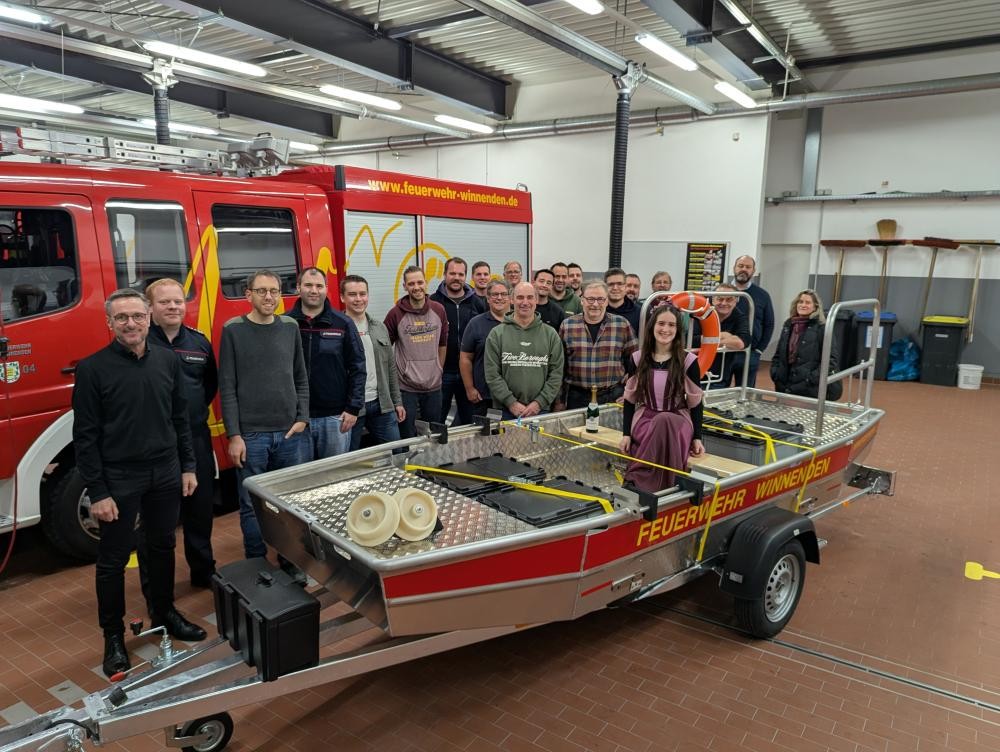 Eine große Gruppe steht hinter einem Boot auf dem "Feuerwehr Winnenden" steht und eine Frau darin sitzt, im Hintergrund sieht man ein Feuerwehrauto.