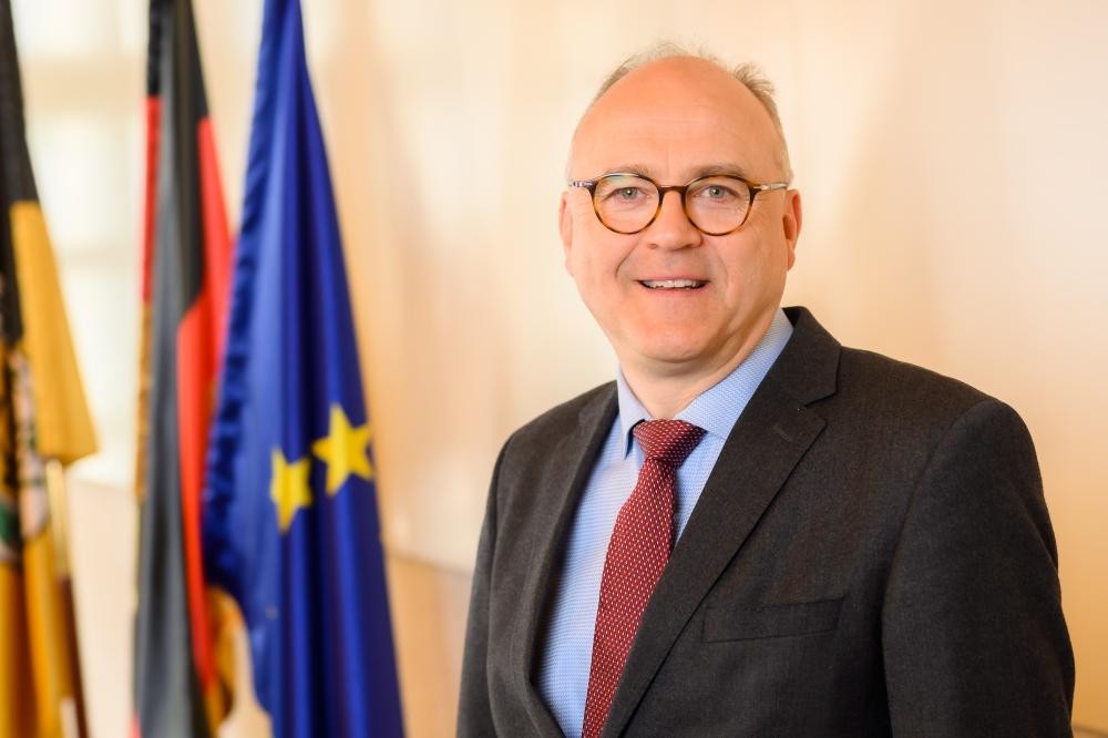 Erster Bürgermeister Norbert Sailer steht in dunklem Anzug, hellblauem Hemd und roter Krawatte vor drei Fahnen.