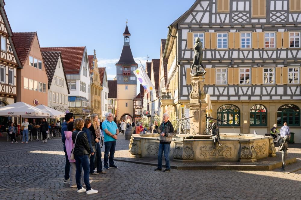 Gruppe von Menschen bei einer Stadtführung vor einem historischen Fachwerkhaus und einem Brunnen mit Statue in einer Altstadt