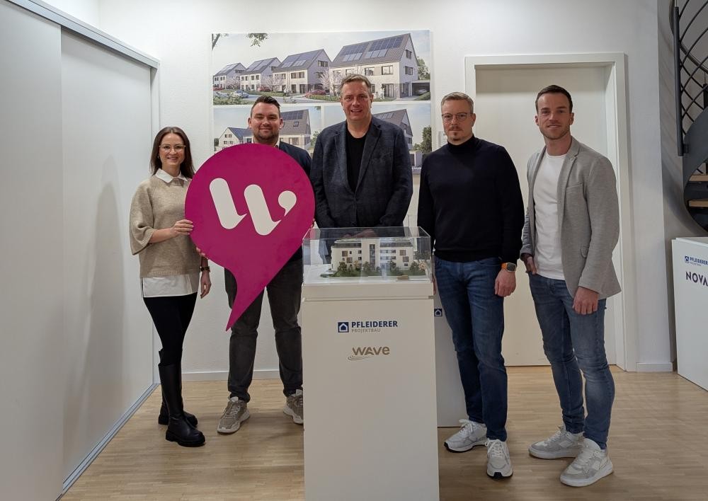 Fünf Personen stehen in einem hellen Raum um einen weißen Sockel mit einem Architekturmodell, eine Person hält ein großes pinkfarbenes Schild mit weißem Logo