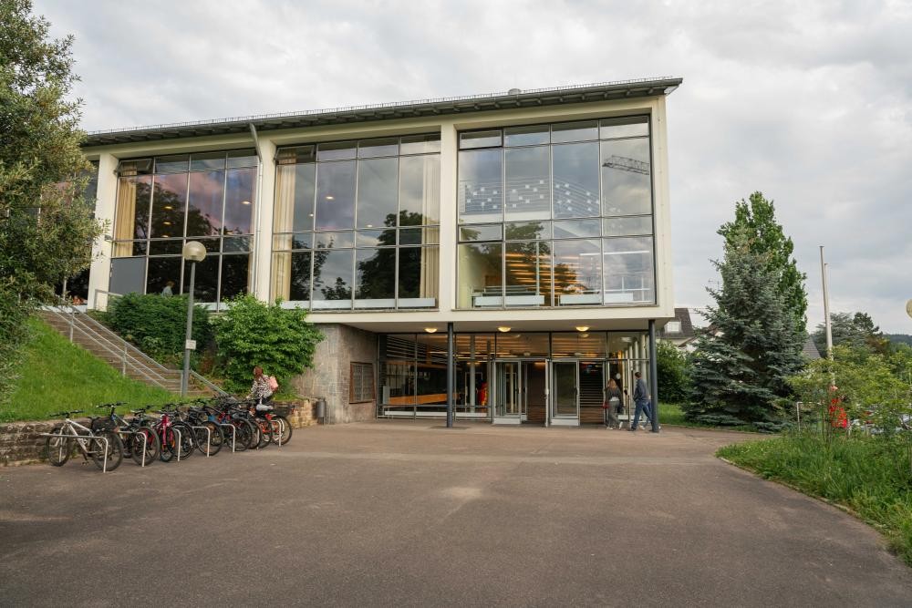 Modernes Gebäude mit großer Glasfassade, davor Fahrradständer und Personen auf einem Weg