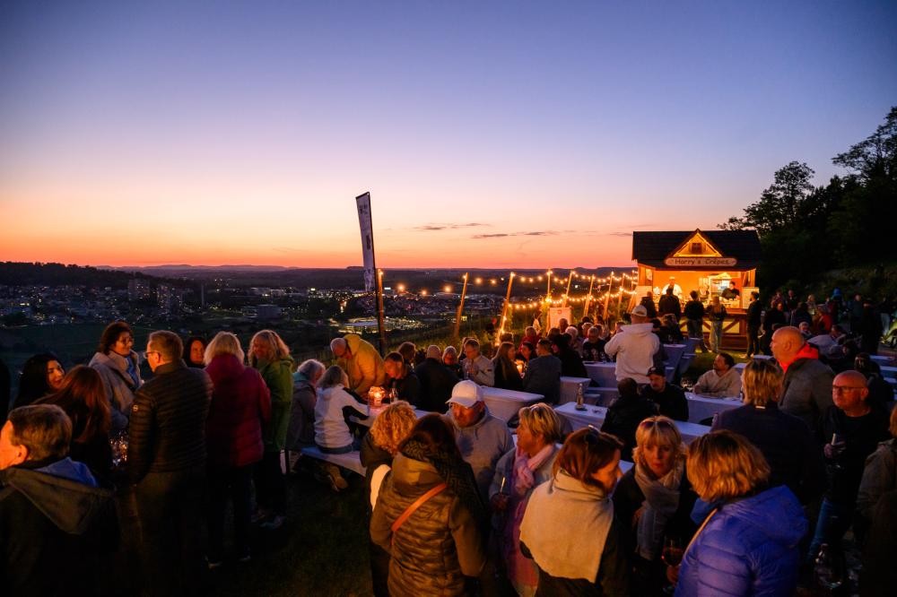 Menschenmenge bei einem Weinfest im Freien am Abend mit beleuchteten Ständen und Blick auf eine Stadt im Hintergrund