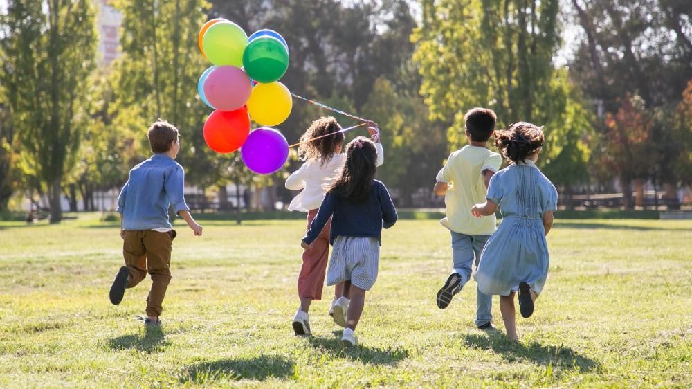 Fünf Kinder laufen auf einer Wiese, eines hält einen Strauß bunter Luftballons