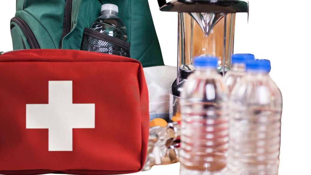 Rotes Erste-Hilfe-Set mit weißem Kreuz neben mehreren Wasserflaschen und einem grünen Rucksack