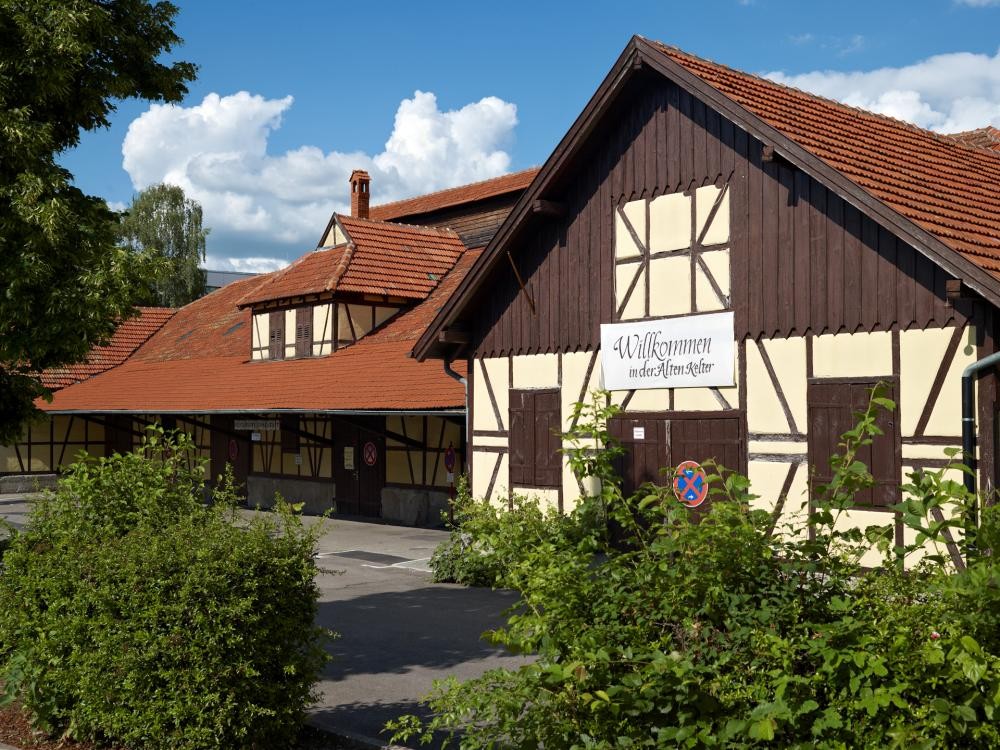 Fachwerkgebäude mit rotem Ziegeldach und Schild mit der Aufschrift 'Willkommen in der Alten Kelter' vor blauem Himmel und grünen Büschen