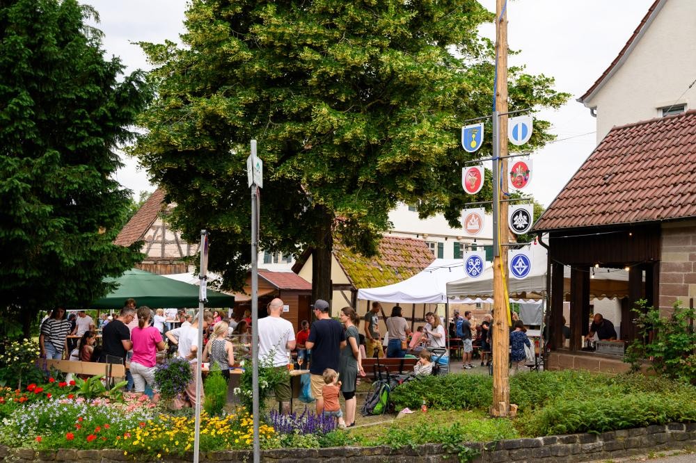 Menschenmenge bei einem Fest mit Ständen und einem Baum mit Wappenschildern an einem Holzpfahl in einem Dorf