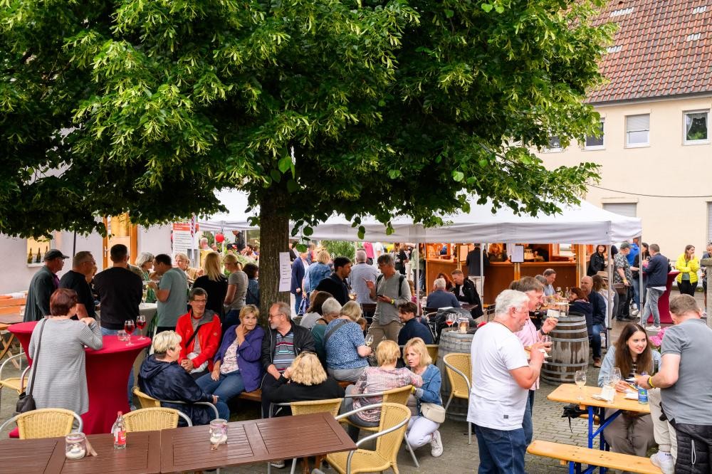 Menschen sitzen und stehen an Tischen und Stehtischen unter einem großen Baum bei einem Straßenfest mit Verkaufsständen im Hintergrund