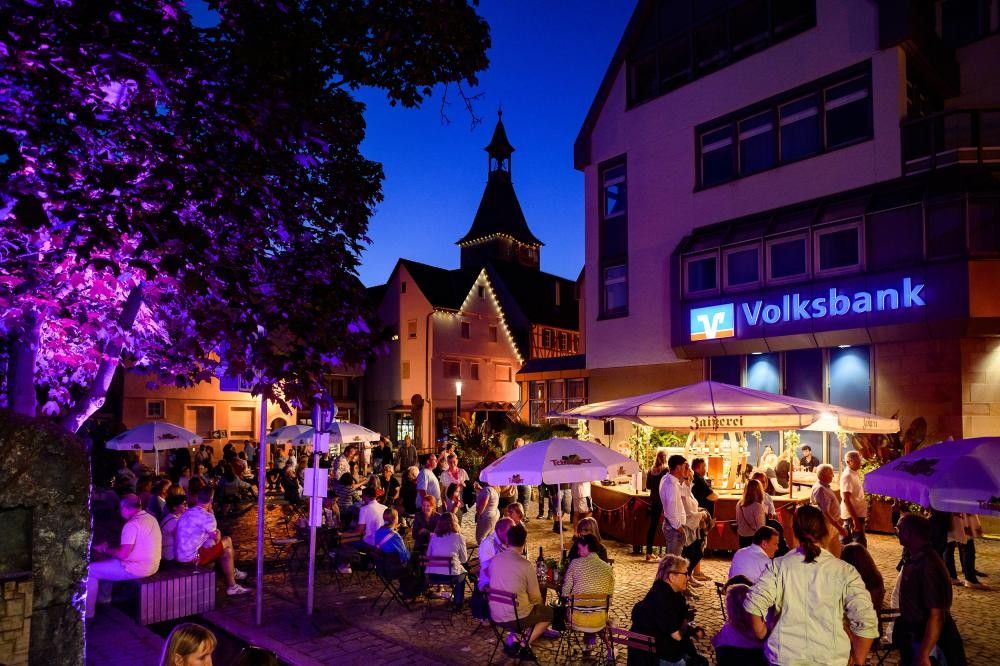 Menschen sitzen und stehen bei einem abendlichen Stadtfest vor einem beleuchteten Volksbank-Gebäude mit Stufendach und Turm im Hintergrund