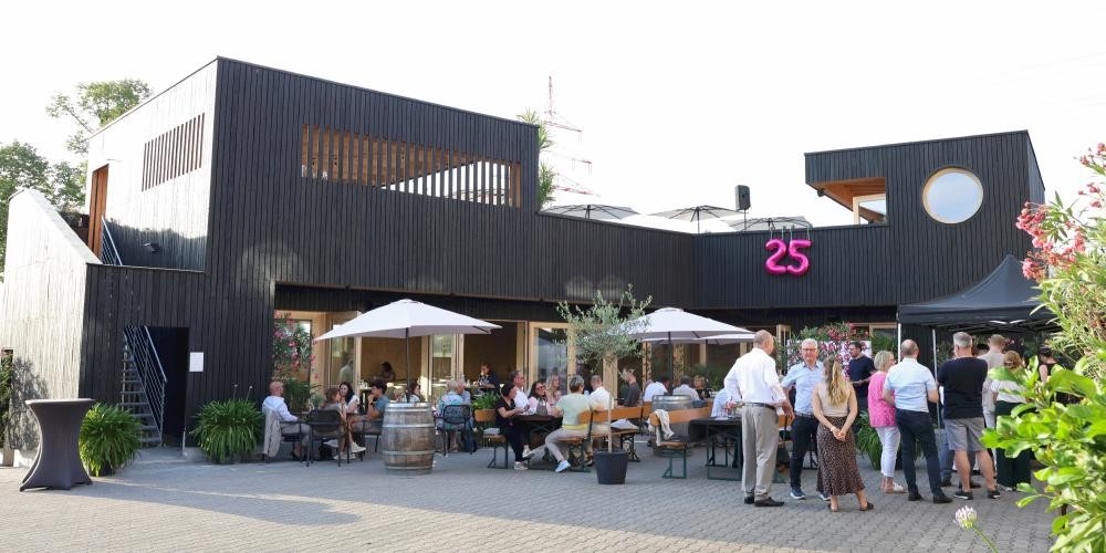 Menschen sitzen und stehen vor einem modernen, zweistöckigen Gebäude mit dunkler Holzfassade und einer pinkfarbenen Leuchtschrift '25'