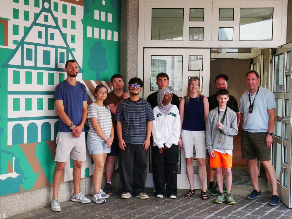 Gruppe von zehn Personen steht vor einer Wand mit buntem geometrischem Muster und Fenstern im Hintergrund