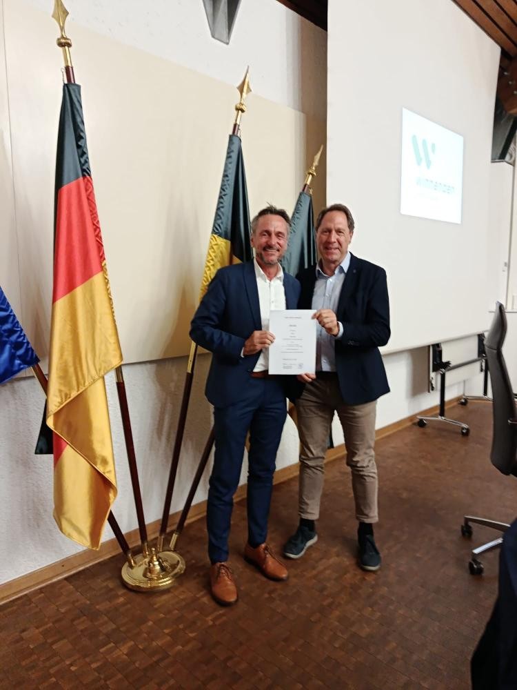 Zwei Männer in Anzügen stehen vor drei Fahnen und halten gemeinsam eine Urkunde in einem Konferenzraum
