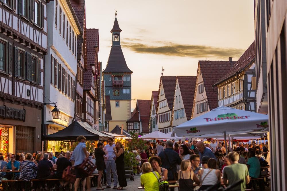 Menschenmenge sitzt und steht in einer Altstadtstraße mit Fachwerkhäusern und einem Turm im Hintergrund bei Abendlicht