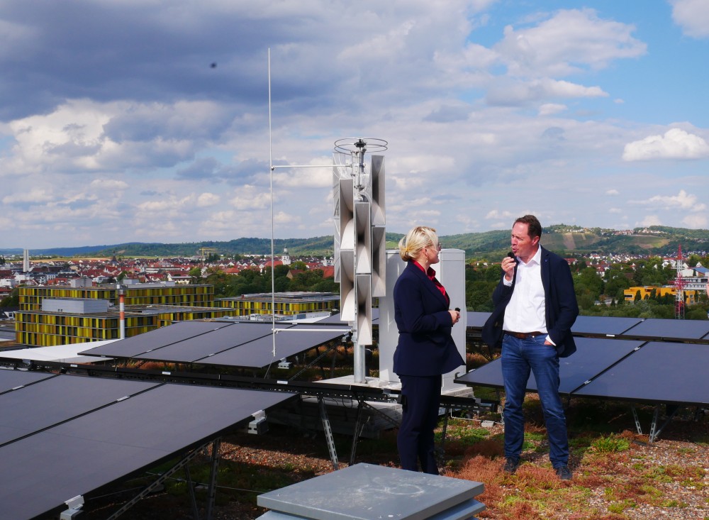 Zwei Personen im Anzug stehen auf einem Dach mit Solarpaneelen und unterhalten sich vor einer Stadtlandschaft unter blauem Himmel mit Wolken