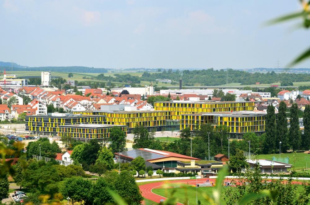 Blick auf ein modernes, mehrstöckiges Gebäude mit gelb-schwarzer Fassade vor einer ländlichen Kulisse mit Feldern und weiteren Gebäuden
