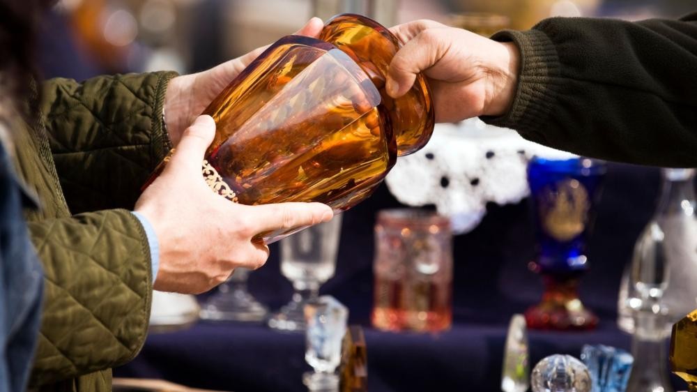Zwei Personen tauschen ein großes bernsteinfarbenes Glasgefäß auf einem Markt mit weiteren Gläsern im Hintergrund