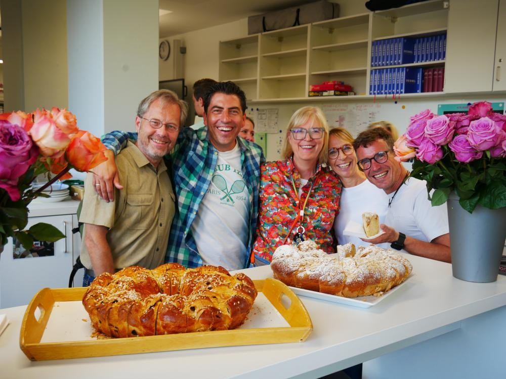 Fünf lächelnde Personen stehen in einer Küche hinter einem Tresen mit zwei großen Hefezöpfen und Blumenvasen mit Rosen.
