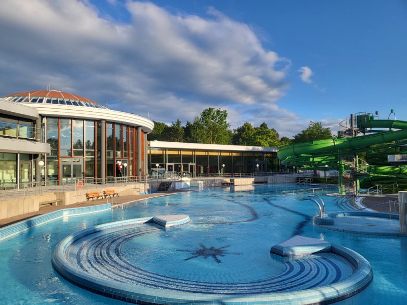 Außenansicht eines Schwimmbads mit rundem Becken, Wasserrutsche und Gebäudekomplex unter blauem Himmel