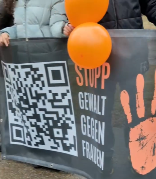Jugendliche halten ein Banner mit der Aufschrift "Stopp Gewalt gegen Frauen" und orangene Luftballons.
