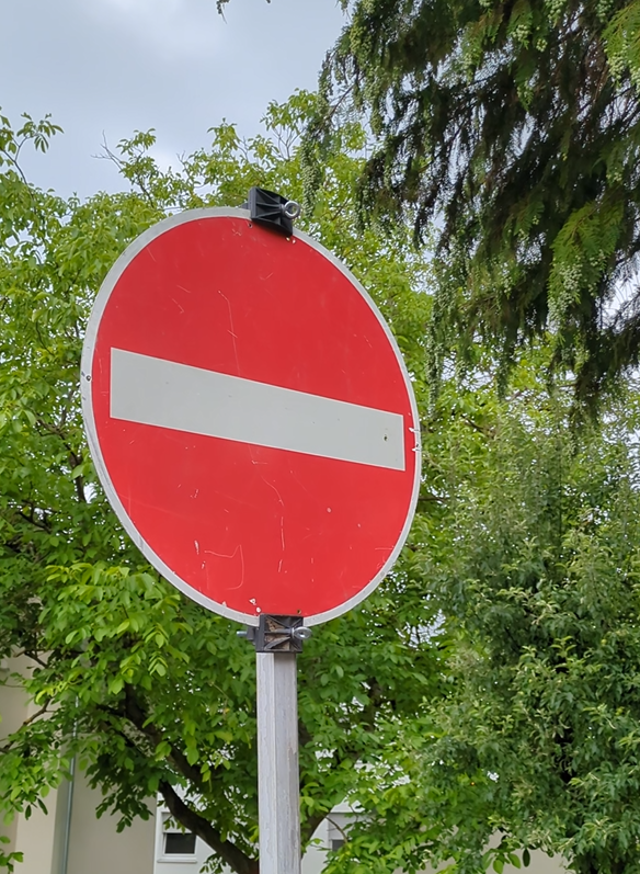 Rotes Verkehrsschild mit weißem Querbalken vor grünen Bäumen und einem Haus im Hintergrund