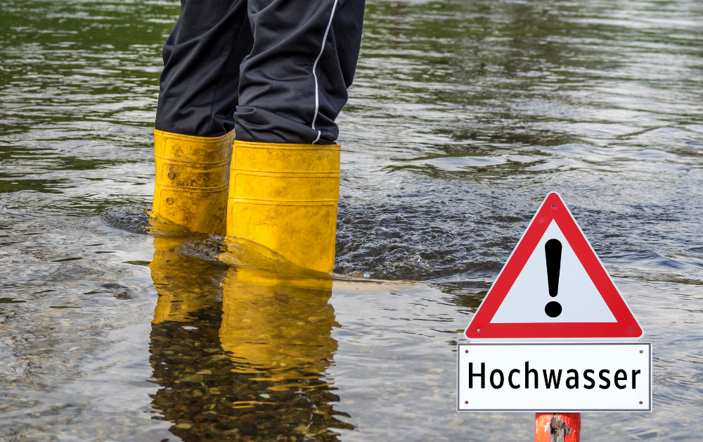 Beine in gelben Gummistiefeln stehen im Hochwasser neben einem Warnschild mit Ausrufezeichen und der Aufschrift 'Hochwasser'