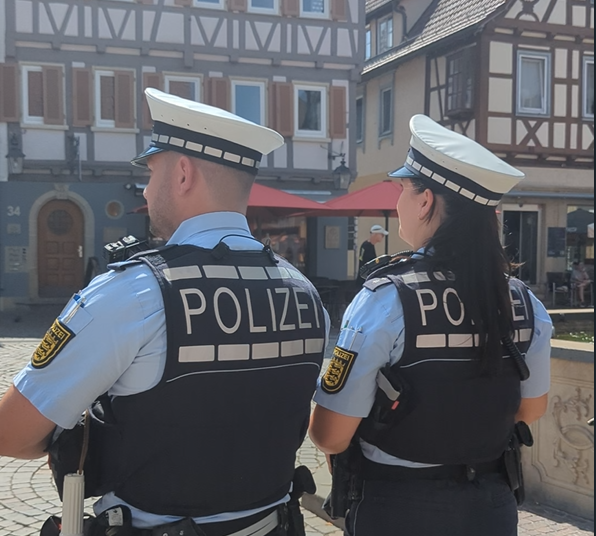 Zwei Polizisten mit weißen Mützen und schwarzen Schutzwesten mit der Aufschrift »POLIZEI« stehen mit dem Rücken zur Kamera vor Fachwerkhäusern
