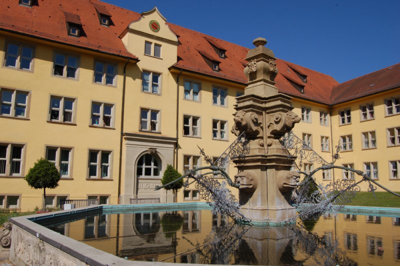 Das Schloss Winnetal mit seiner gelben Fassade und dem Roten Satteldach im Hintergrund. Davor der Brunnen mit detailreichen Verzierungen.