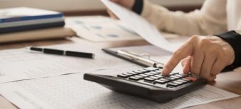 Symbolbild: Person tippt in einen Taschenrechner auf einem Holztisch