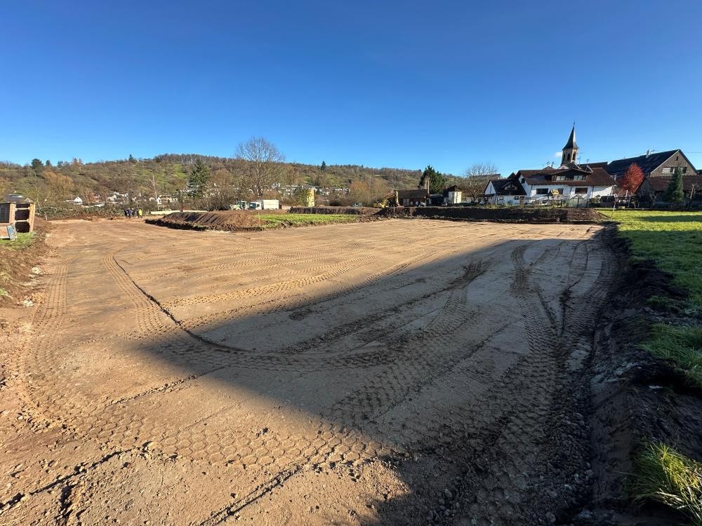 Großflächig planiertes, unbefestigtes Erdgelände mit Spuren von Baumaschinen vor einer Dorfkulisse mit Kirche und Häusern