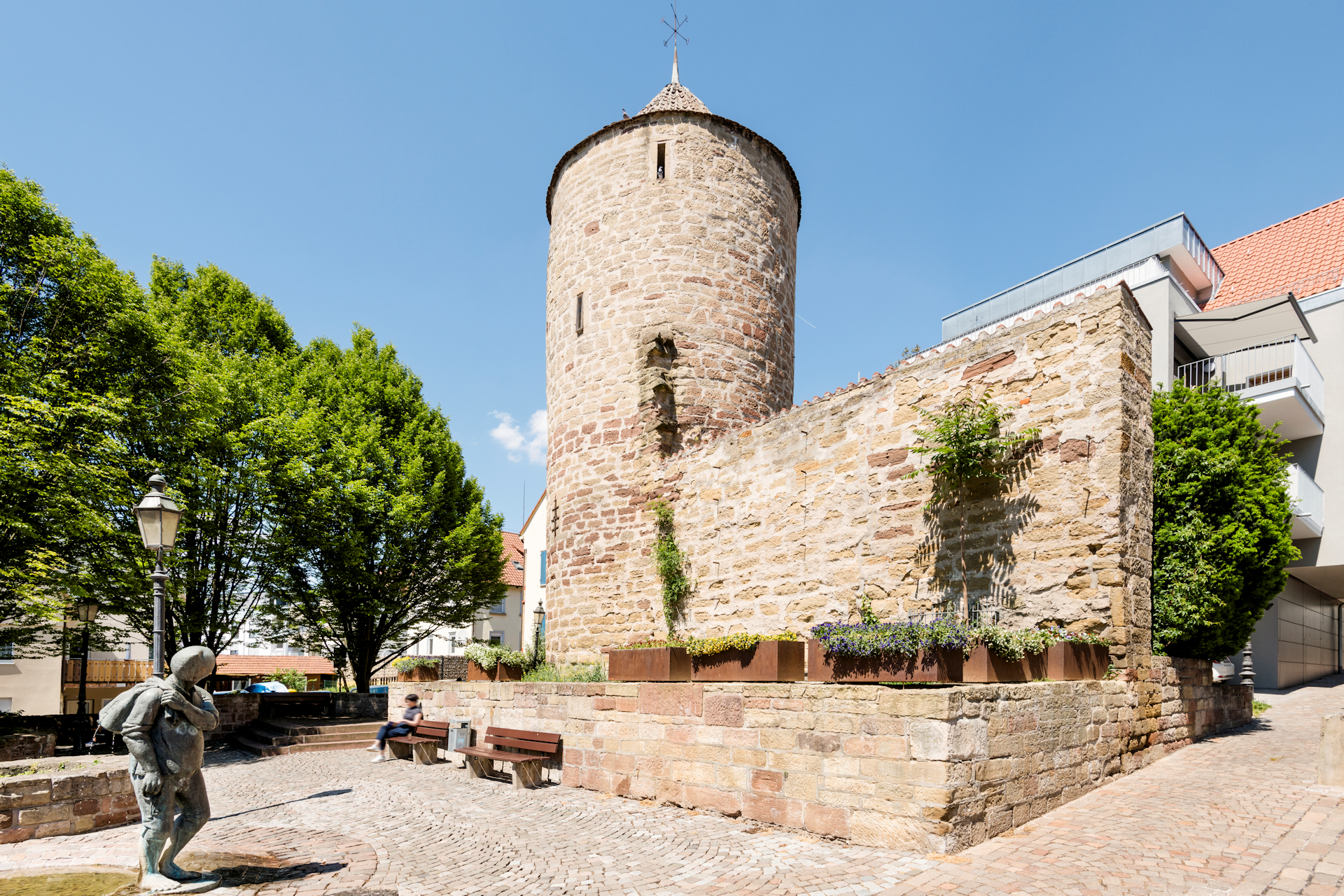 Der Diebsturm aus hellen Steinen gemauert bei strahlen blauem Himmel. Davor der Diebsbrunnen mit einer Brunnen Figur.