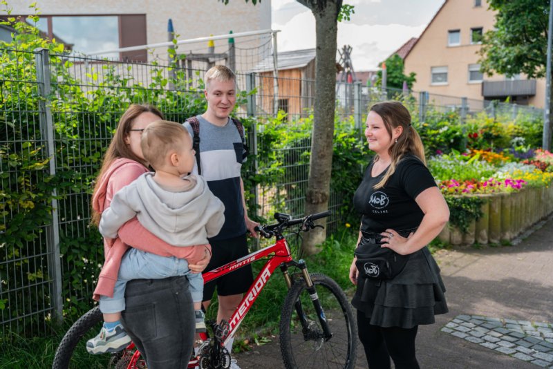 Drei junge Menschen stehen zusammen. Eine der jugen Frauen hat ein Kleinkind auf dem Arm. Die andere träge ein Oberteil der Mob´s-Mitarbeitergruppe