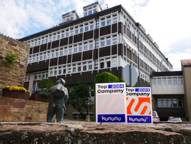 Im Vordergrund stehen zwei Auszeichnungen von Kununu für die TOP-Company 2024 und 2023. Im Hintergrund ist das Rathaus zu sehen. 