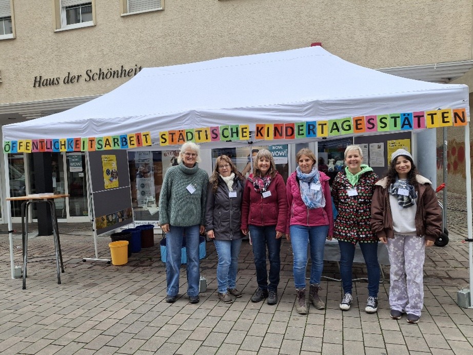 Sechs Frauen stehen vor einem weißen Pavillon mit bunter Buchstabenkette und dem Schriftzug 'Haus der Schönheit' an der Hauswand