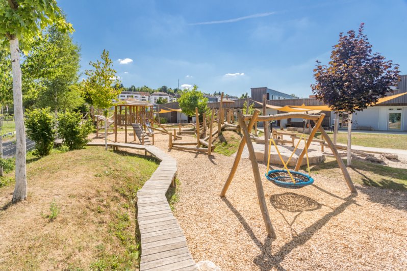 Mehrere Spielgeräte wie eine Nestschaukel und ein Sandkasten vor einer Kita.