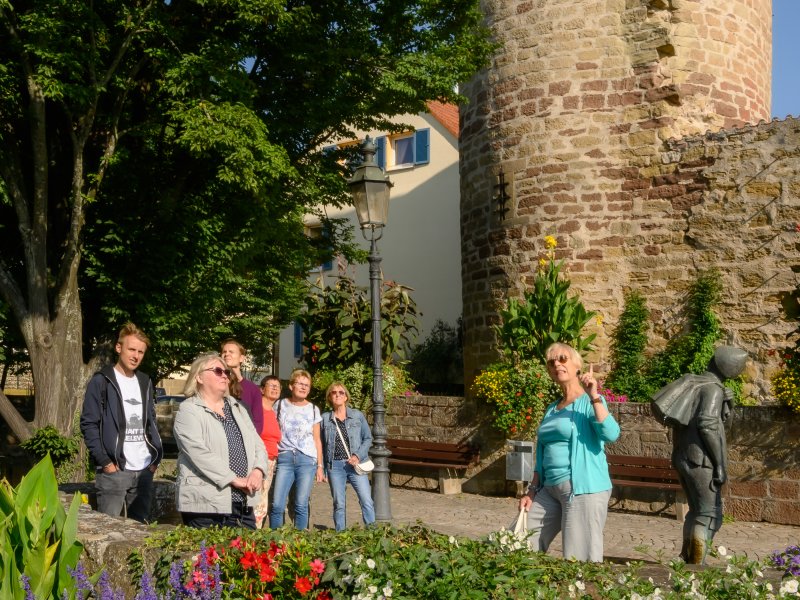 Eine Gruppe Menschen steht beim Diebsturm um die Stadtführerin, die erzählt und deutet auf die gegenüberliegende Kirche