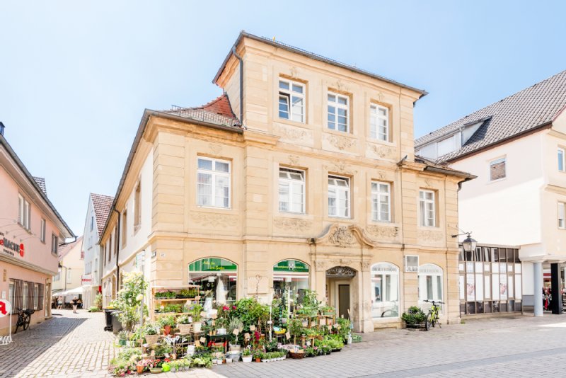 Das Haus Sator in der Marktstraße ist beige mit großen Fenstern. Vor dem Haus stehen Blumen und Gestecke für den Gartwen