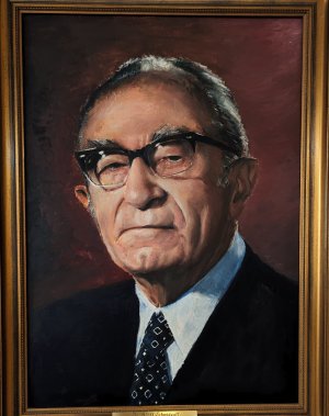 Das Ehrenbürgergemälde zeigt Dr. Adolf Schmidgall im dunklen Anzug mit gemusterter Krawatte und dunkler Brille ernst den Betrachter anschauend.