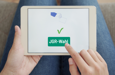 Symbolbild: Online Wahl; Finger tippt auf IPad, dort ist ein Haken sowie die Aufschrift "JGR-Wahl" zu sehen.