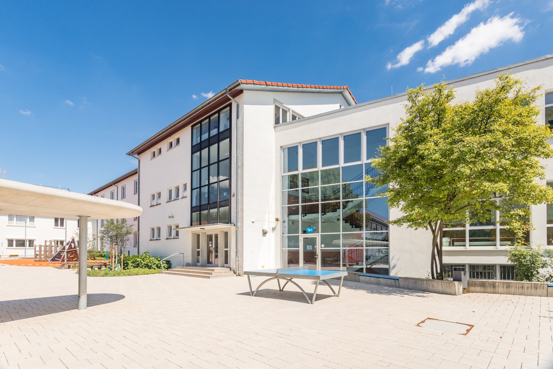 Auf dem Schulhof der Geschwister-Scholl-Realschule stehen eine Tischtennisplatte, ein großer Baum, eine Überdachung und im Hintergrund ein Klettergerüst.