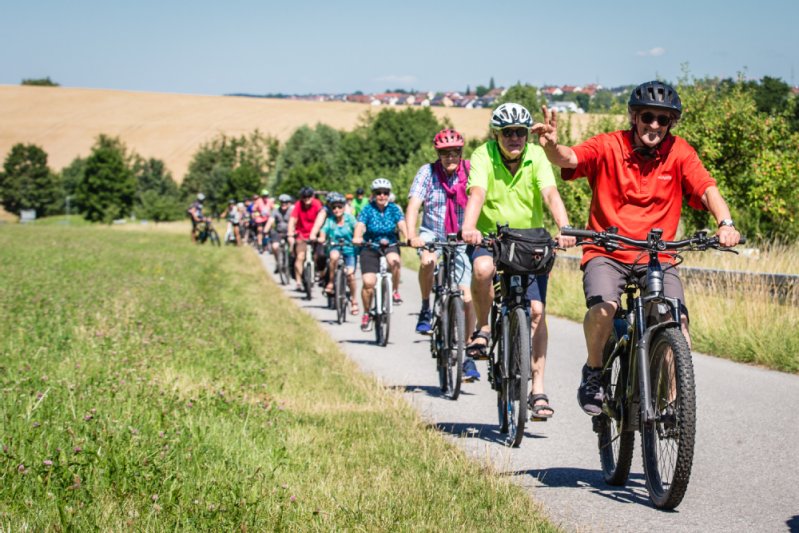 Radfahrer fahren zusammen an einem Feld entlang. Ein Radfahrer posiert dabei vergnügt für die Kamera