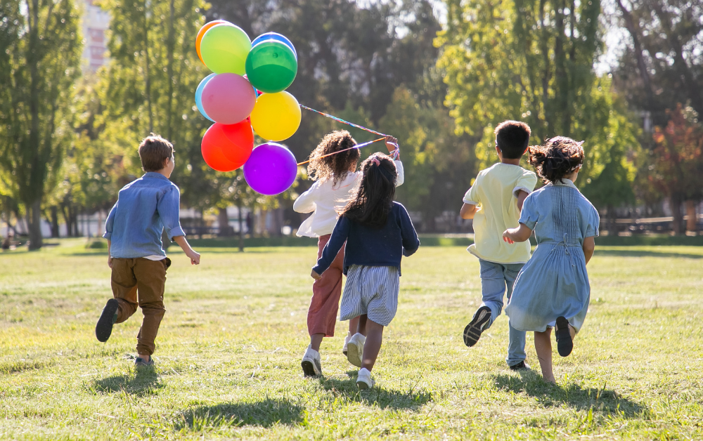 Fünf Kinder laufen auf einer Wiese davon, eines hält einen Strauß bunter Luftballons
