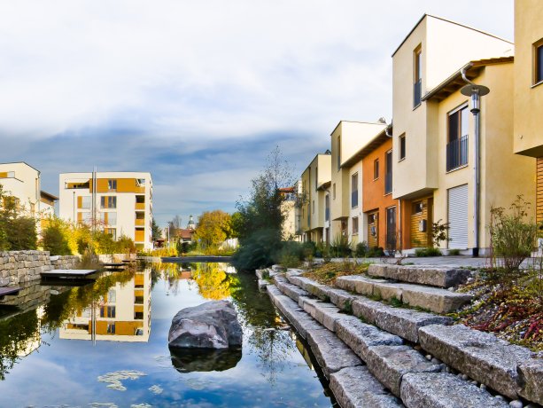 Das Wohngebiet Adelsbach mit dem See in der Mitte und den darumstehenden Häusern in Beige und Gelb.