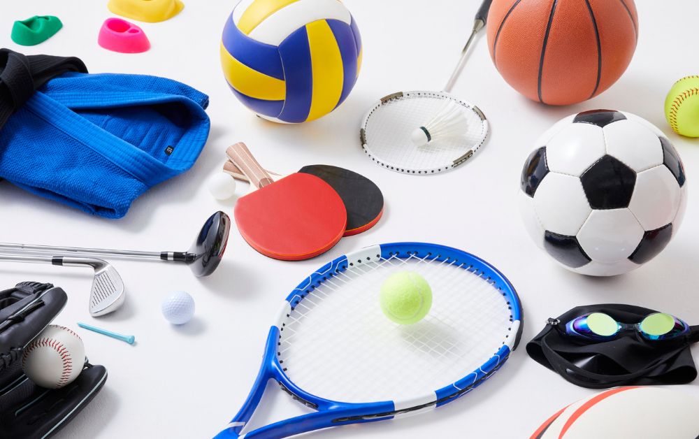 Verschiedene Sportgeräte wie Volleyball, Basketball, Fußball, Tennisracket mit Ball, Tischtennisschläger, Badmintonschläger mit Federball, Baseballhandschuh mit Ball, Golfball und Golfclub, Schwimmbrille und Judoanzug auf weißem Untergrund