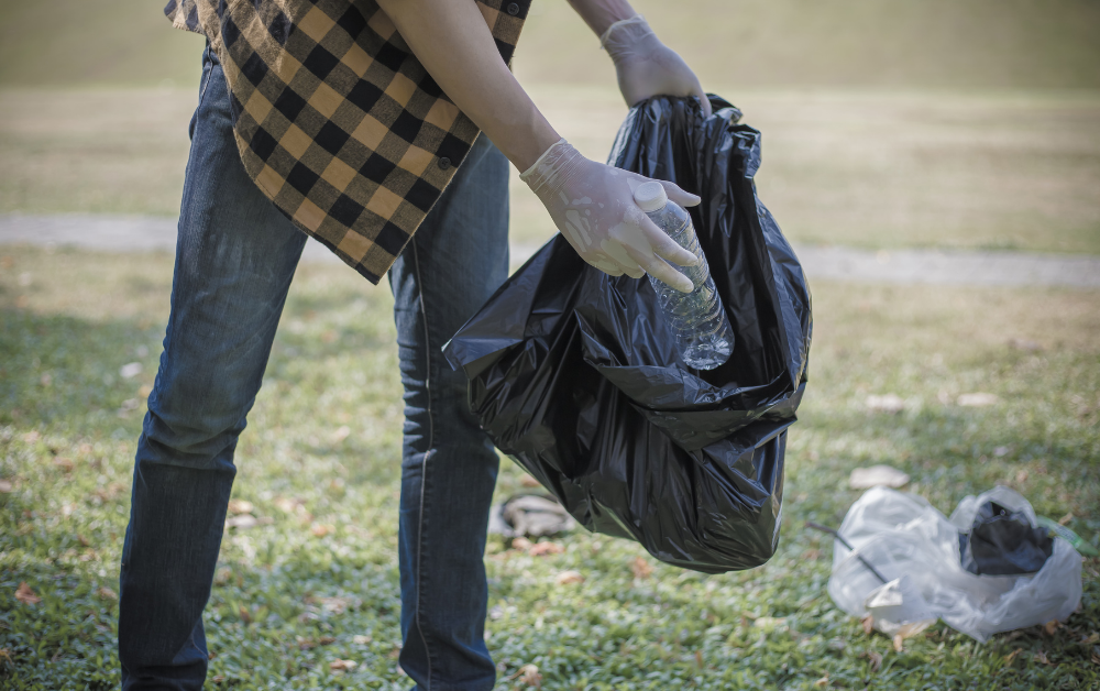 Person mit Handschuhen hält eine schwarze Mülltüte und entsorgt eine Plastikflasche auf einer Wiese.